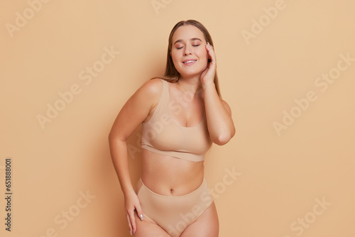 Pretty model dressed in beige underwear posing with closed eyes on background holding one hand on her face, body comfort concept, copy space
