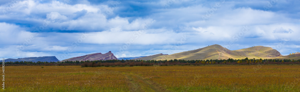 Panoramic landscape with Khakassia steppe at early autumn day in Siberia, Russia. Mountain range Sunduki on the horizon under blue sky. Overgrown dirt road in a field