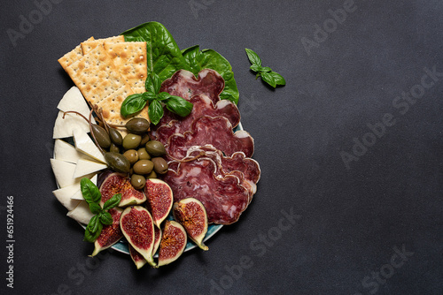 Antipasti set with figs, cheese, jamon, olives, capers and crackers on the plate on black stone background top view.