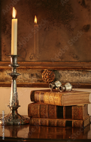 Antique leather-bound books next to a silver candelabra. In the background is an antique Venetian mirror with antique opera glasses resting on books. Stock Image.