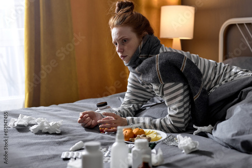 serious redhead woman with cold blowing runny nose and swollen throat sitting on bed at home. female in bedroom, with medicaments pills. sick female in domestic interior, having temperature high fever