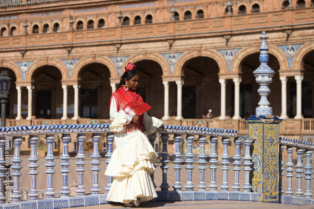 Young black woman dressed as a flamenco gypsy in a famous square in Seville, Spain. She wears a beige dress with ruffles and red shawl and red fan. Flamenco cultural heritage of humanity.