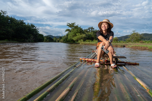 Female tourist carries a backpack, travels on a bamboo raft, smiles happily, relaxes on vacation, river rafting, travel, nature, forest, mountains, both feet are wet on the bamboo raft. © Supavadee