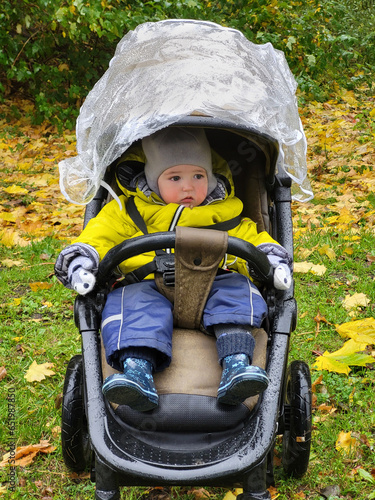 Child in a baby stroller in an autumn park with yellow foliage during the rain