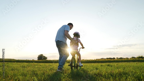 Loving father teaches toddler daughter training bicycle riding in sunset field