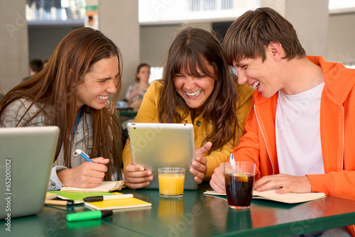 Three smiling Caucasian students snacking drinks and doing homework using tablet in university cafeteria. Young people having fun looking at web on device during recess. Generation z and technologies.