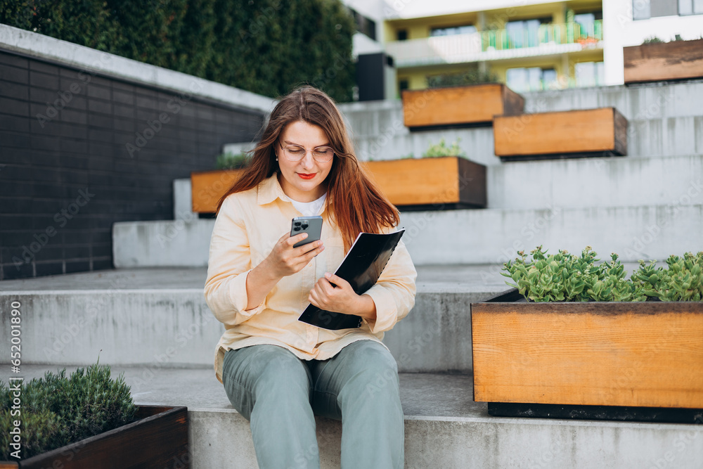 Elegant young 30s woman formally dressed, sitting on the bench and holding a folder with documents outdoors. Businesswoman with smart phone standing at the city.