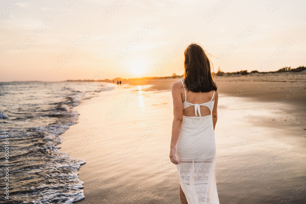 beautiful woman walking along the beach shore with sunset light dressed in white