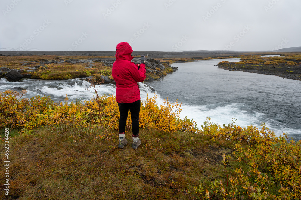 Woman in red raincoat photographing Grafarlandsfoss falls in northern Icelandic highlands amidst colorful autumn vegetation.
