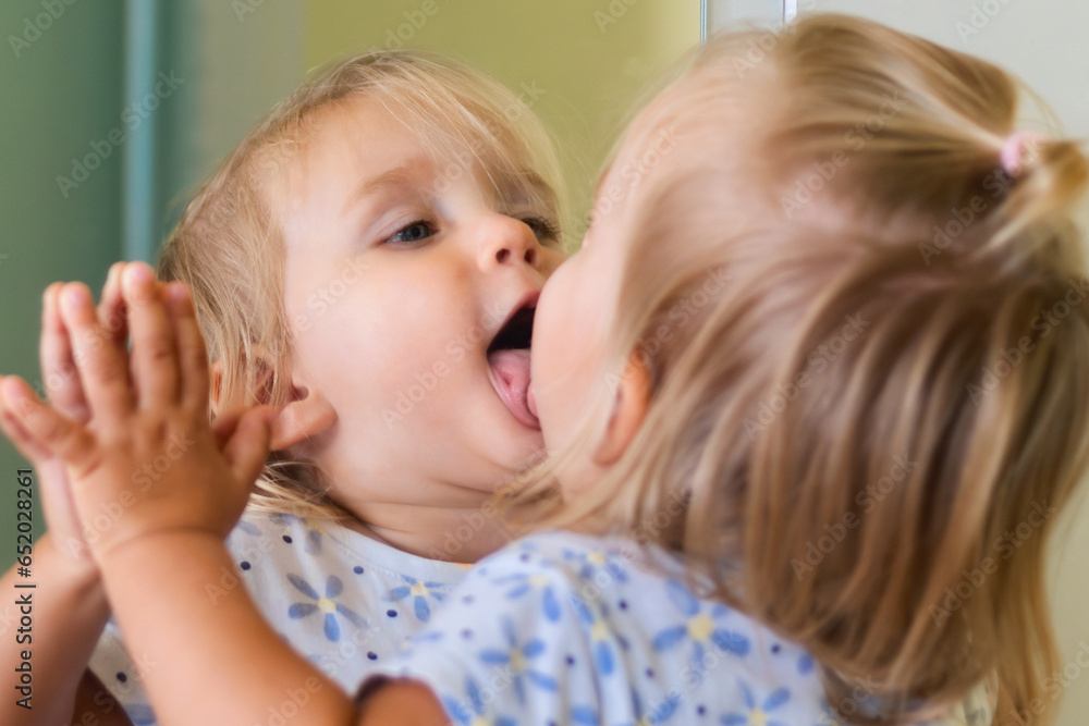 Funny little child blonde girl makes faces, shows tongue in the mirror Funny little child blonde girl makes faces, shows tongue in the mirror