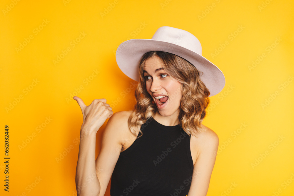 Attractive blonde curly woman wear white hat and black top smiling happy pointing with hand and finger to the side standing isolated on yellow background. Wow emotions, sale here, attention! Attractive blonde curly woman wear white hat and black top smiling happy pointing with hand and finger to the side standing isolated on yellow background. Wow emotions, sale here, attention!