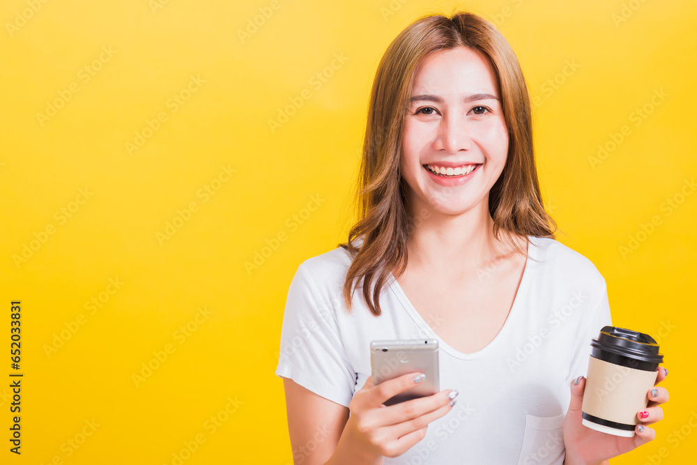 Portrait Asian Thai beautiful happy young woman standing smile, using mobile phone her holding coffee paper cup, looking to camera, studio shot isolated on yellow background, with copy space