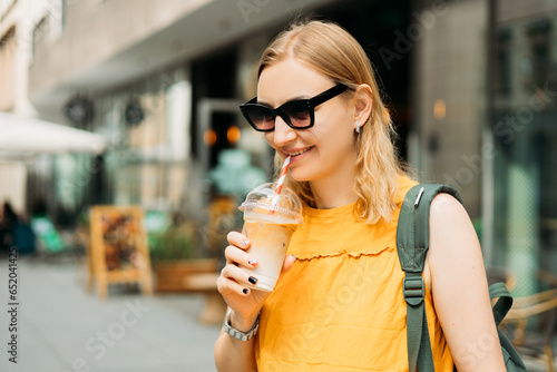 Beautiful fashionable young woman in sunglasses with backpack standing on city street. Urban lifestyle concept. Attractive female drinking a ice coffee in plastic cup.
