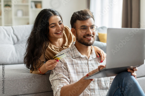 Indian couple with laptop computer shopping online from home, paying for order in internet, purchasing and buying things, sitting in living room