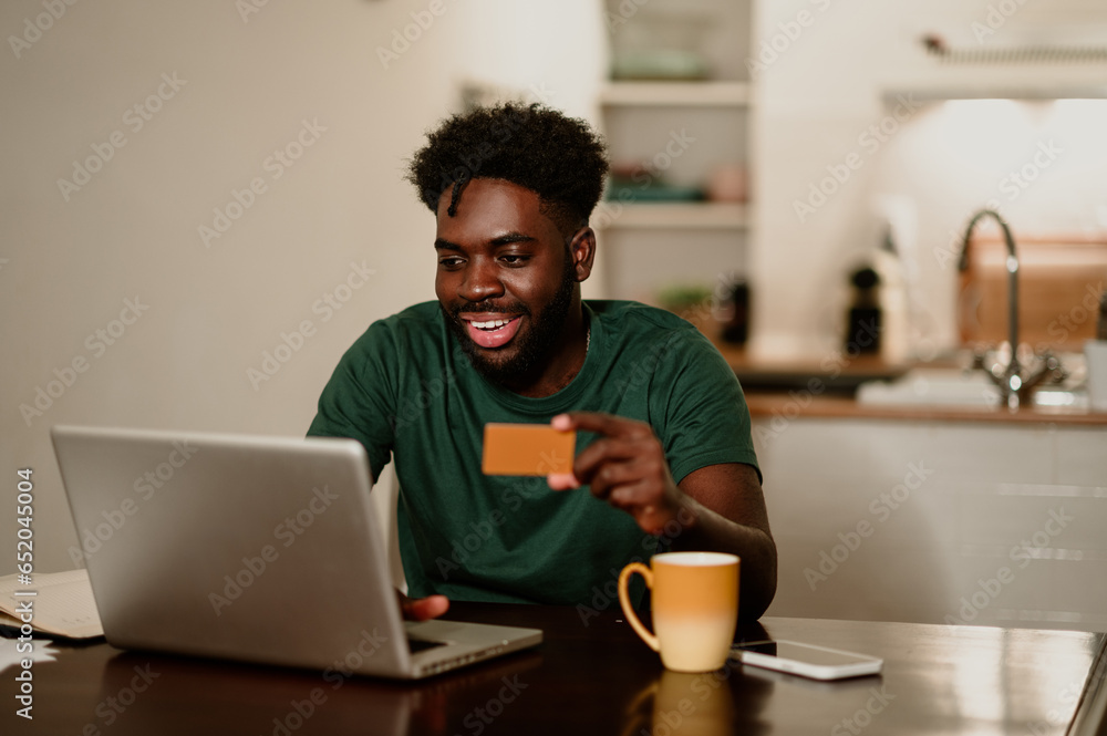 Portrait of a happy african american man sitting at home and using a credit card and a laptop for online shopping. Portrait of a happy african american man sitting at home and using a credit card and a laptop for online shopping.