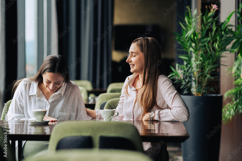 Two young women are sitting together in a coffee shop, chatting, drinking coffee, and having a good time.