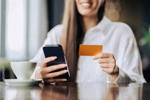 Close up of a young woman holding a credit card and her phone, and using e-banking, and online banking apps.