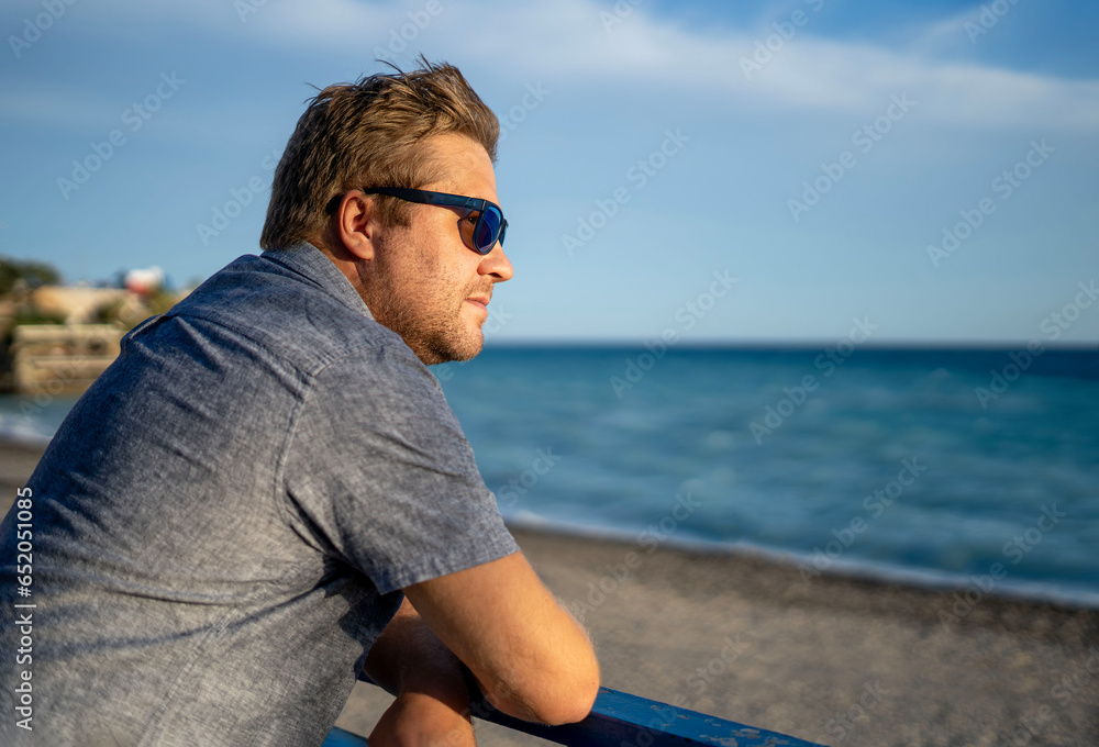 A blond man in sunglasses looks into the distance at the Mediterranean Sea.
