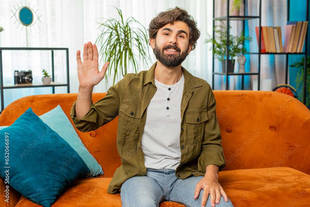 Fototapeta premium Hello. Middle eastern handsome man smiling friendly at camera and waving hands gesturing hello hi, greeting or goodbye, welcoming with hospitable expression at home. Guy sitting on sofa in living room