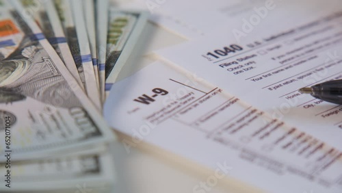 American dollar tax bill, pen calculator and U.S. money. 1040 and W9 individual tax form are placed on desk of American citizen. rotate camera shot photo