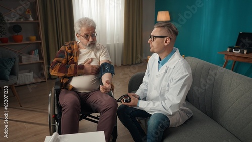 Full-size shot of a doctor checking up his patient at home. He is using a sphygmomanometer, a device for measuring blood pressure of an eldery man on a wheelchair. photo