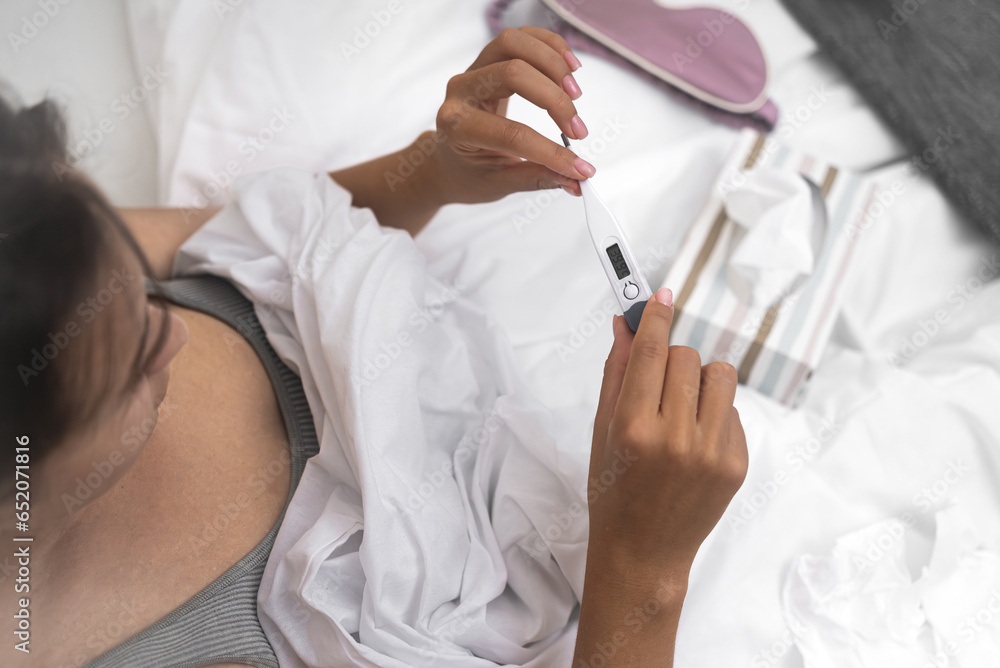 Naklejka premium A woman, ill and nestled in her bed, gauges her body temperature using a thermometer, tissues nearby, contending with fever and sickness. 