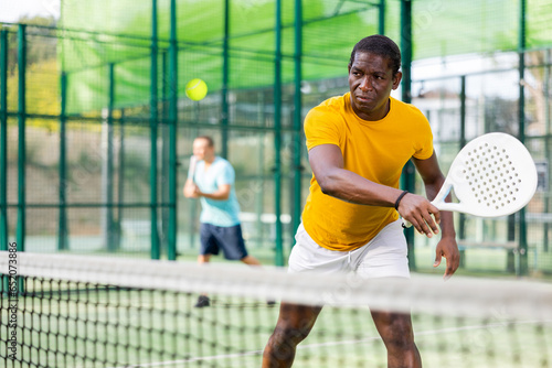 African american male player ready to hits the ball while playing padel on a hard court © JackF