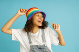 Pretty smiling woman in rainbow cap dancing with raised hands standing over blue studio background
