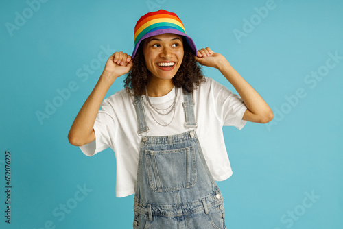 Smiling woman puts rainbow cap while looks away on blue studio background