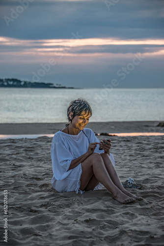 Happy, mature woman sitting on the sand on the beach sending a message on her cell phone with the sea behind her., in Punta del Este, Uruguay. photo