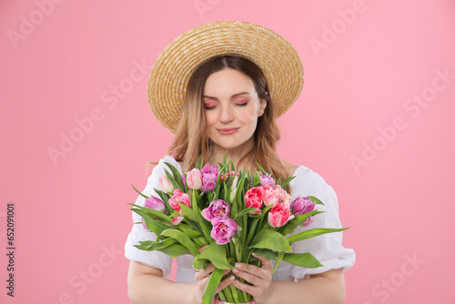 Happy young woman in straw hat holding bouquet of beautiful tulips on pink background