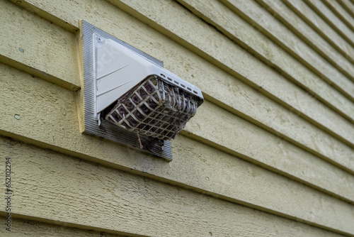 Fotografija The exterior of a bright yellow house wall with a white dryer vent exposes the air vent which is clogged with hair, wood, and dirt in the mesh causing a fire hazard