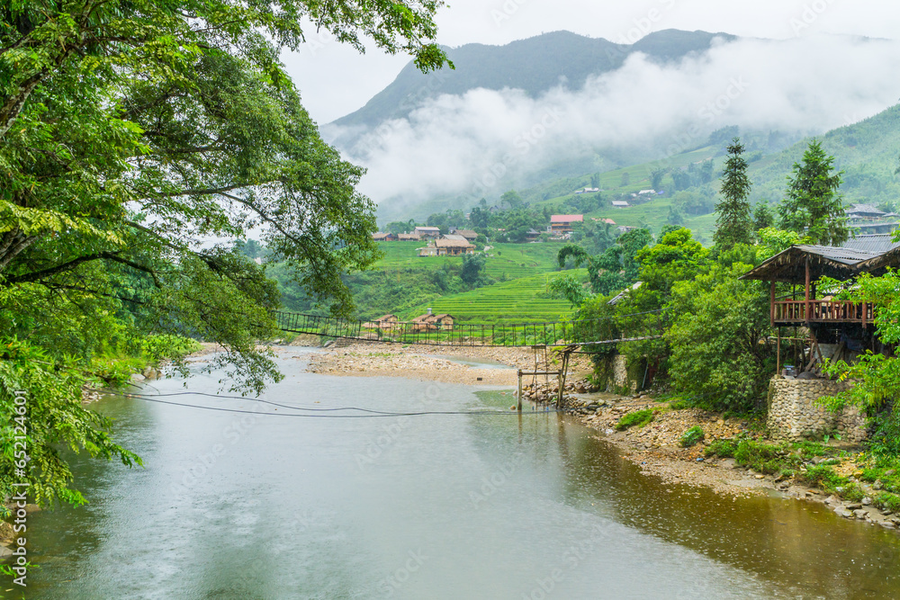 Fototapeta premium A narrow rope bridge over a river in front of a mountain village amongst rice fields at Ta Van in Vietnam