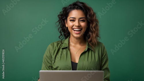Young happy Latin woman winner holding a laptop isolated on a green background. Excited euphoric female model using computer winning online celebrating new great job offer with yes gesture concept.