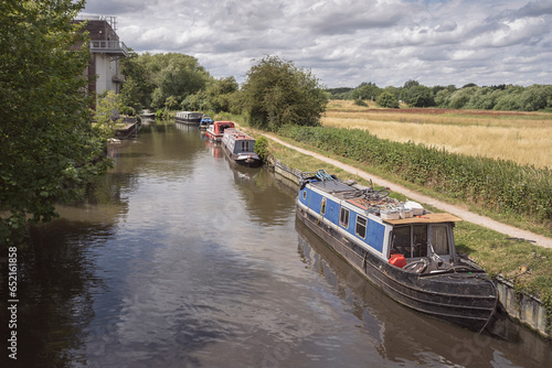 six narrowboats line one side of the river stort near Harlow England among beautiful trees and farmland on a sunny day with a cloudy sky background