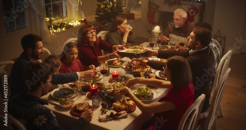 Diverse Group of People, Young and Old, Share a Joyful Christmas Dinner at Home. Family Sitting Behind a Big Dining Table Full of Delicious Food with Roast Turkey Feast. High Angle Evening Footage photo