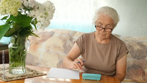 Senior mature elderly gray haired woman with glasses is sitting at table in living room, holding paper bill, using calculator, managing accounts finance, calculating money budget tax