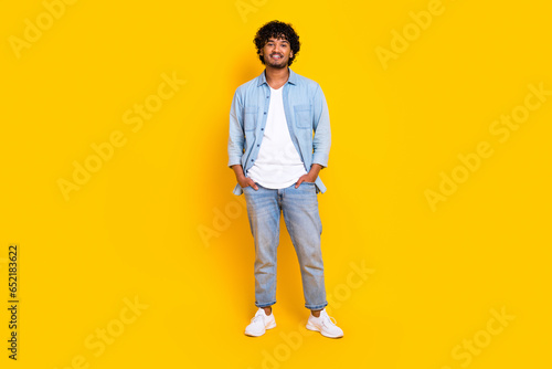 Full size photo of satisfied cool man with curly hairdo dressed denim shirt jeans hold arms in pockets isolated on yellow color background