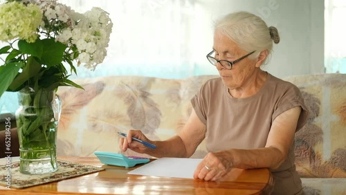 Senior mature elderly gray haired woman with glasses holding paper bill, using calculator, managing accounts finance, calculating money budget tax, is sitting at table in living room.