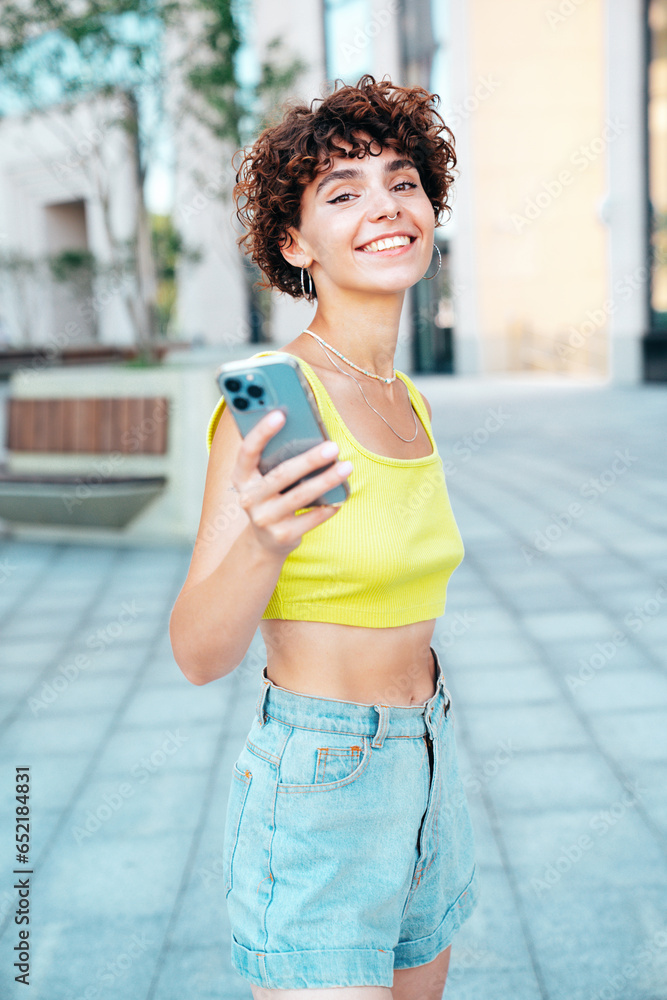 Young beautiful smiling hipster woman in trendy summer clothes. Carefree woman with curls hairstyle, in the street. Positive model holds mobile phone, looks at cellphone screen, uses smartphone apps