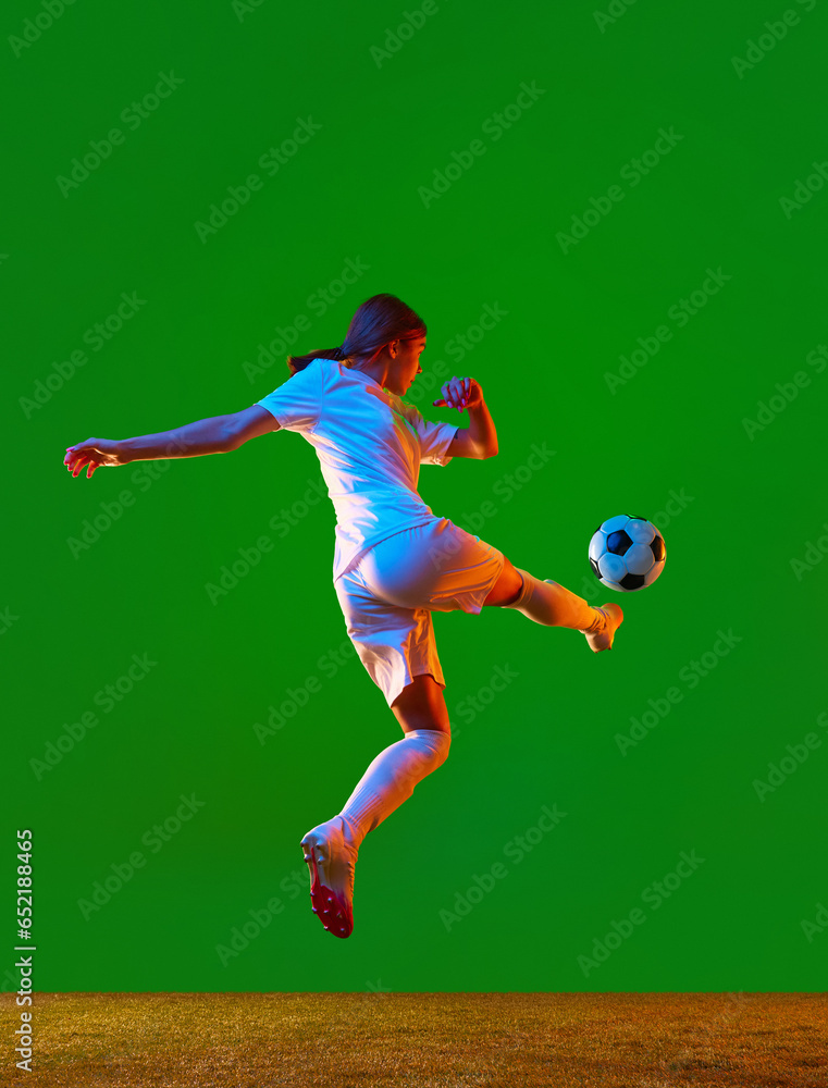 Full-length dynamic image of young girl, football athlete in motion, kicking ball against green studio background in neon light