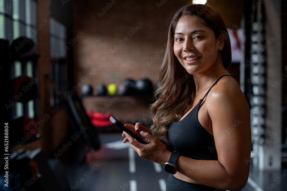 Cheerful sporty woman holding phone, smiling and looking at camera after fitness workout in fitness gym