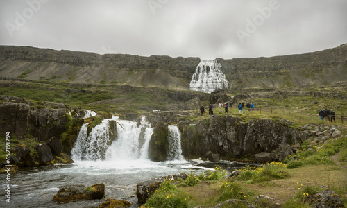 tiny tourists climbing the trail towards the bottom of the Dyndanji waterfall in Westfjords Iceland with Baejarfoss waterfall in the foreground and a cloudy sky background photo
