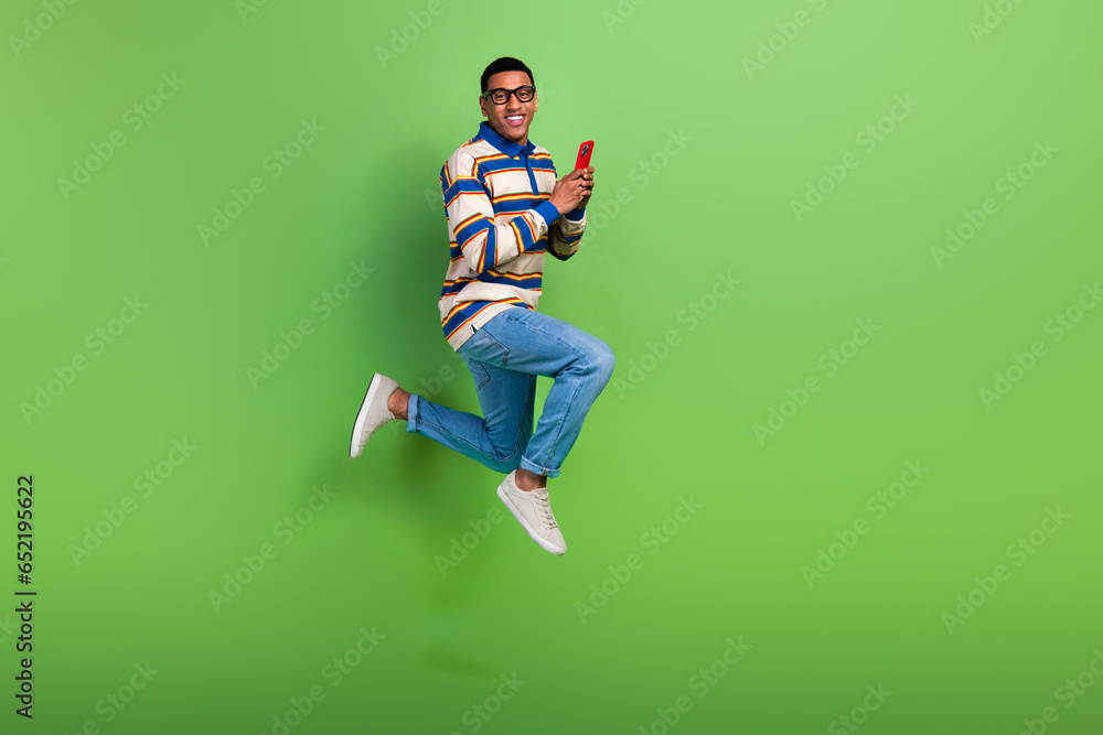 Full size body photo of crazy activity jumping guy influencer with smartphone wear jeans browsing phone isolated on green color background