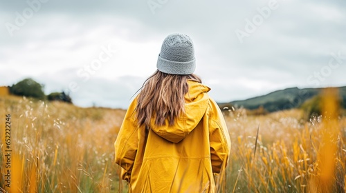 Back view of a girl walking Female in high grass wearing a yellow raincoat and looking away from the camera - Moody fall scenery with a young girl in bright clothing walking in high grass outdoors © BOMB8