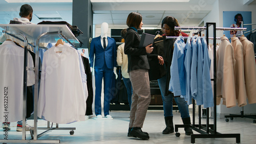 Young employee helping woman to choose shirts, using tablet to check clothes stock in shopping center shop. Store assistant showing new formal or casual wear collection to client.