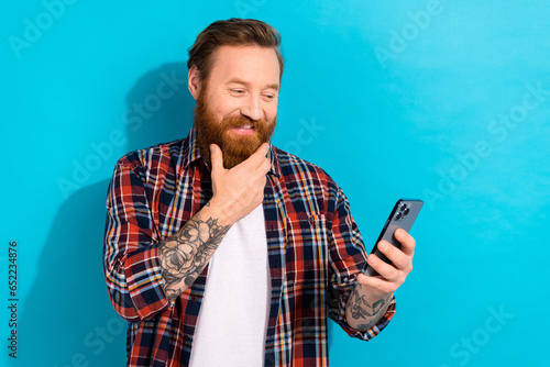 Photo of handsome man with stylish beard dressed checkered shirt look at smartphone hand on chin isolated on blue color background © deagreez