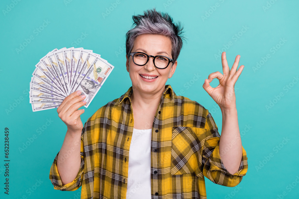 Photo portrait of charming grandmother hold money fan okey symbol wear trendy plaid yellow outfit isolated on cyan color background Photo portrait of charming grandmother hold money fan okey symbol wear trendy plaid yellow outfit isolated on cyan color background