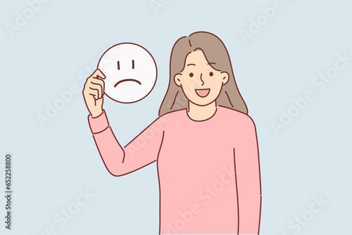 Happy woman holds sad emoticon calling to be optimist and have positive mood for achieve goals.