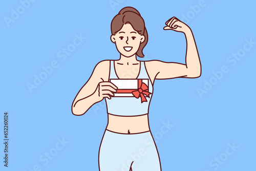 Woman holds gift certificate for fitness club and shows biceps, urging you to do sport. Smiling girl demonstrates fitness voucher with red ribbon presented for birthday and motivating to visit gym